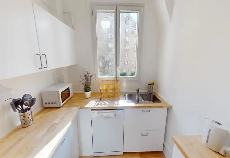 Compact, bright kitchen with wooden countertops, built-in sink, dishwasher and microwave. Natural light from the central window highlights tidy work surfaces and simple, modern storage.