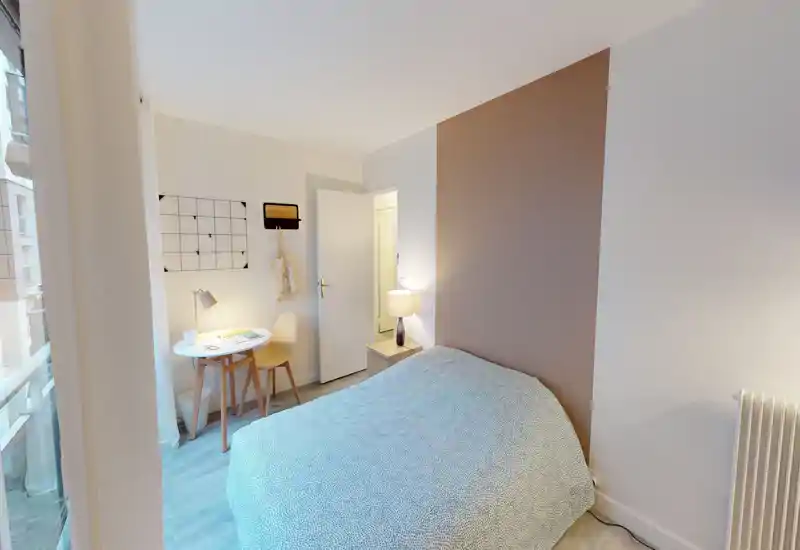 Camera da letto luminosa e accogliente con letto matrimoniale, lampada da comodino e un piccolo tavolo da lavoro vicino alla finestra — arredi moderni e pavimento effetto legno.