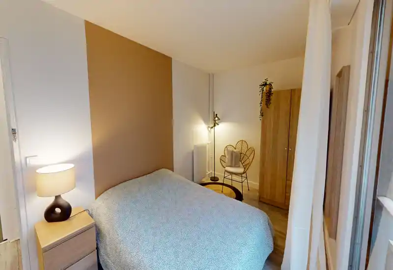 Camera da letto luminosa e accogliente con letto matrimoniale davanti a una parete d'accento calda, comodino con lampada e tessuti morbidi; una sedia in rattan e un armadio completano l'arredamento.