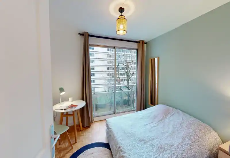Camera da letto luminosa e ordinata con letto matrimoniale, lampada da comodino e piccolo angolo studio vicino a una grande finestra scorrevole che garantisce luce naturale.
