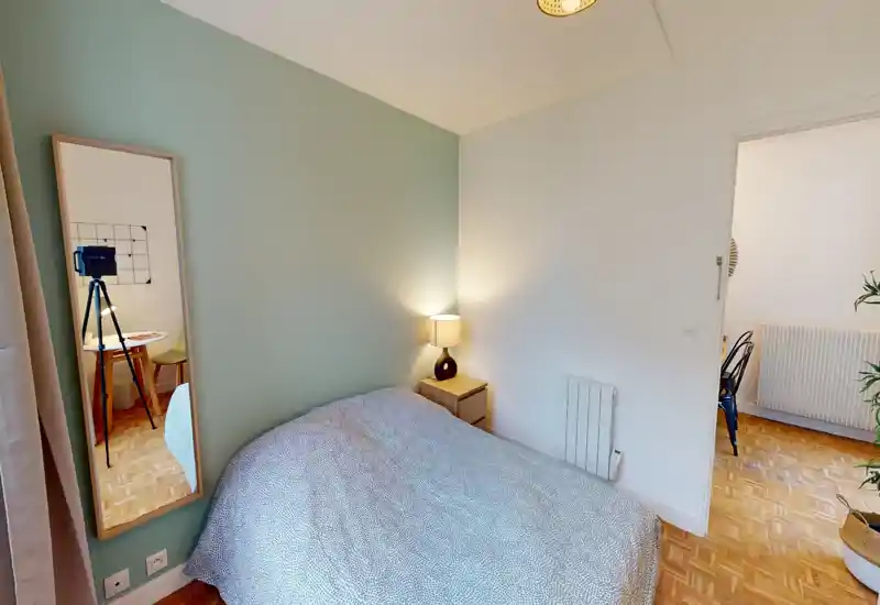 Camera da letto luminosa e ordinata con un letto matrimoniale contro una parete verde, comodino con lampada e uno specchio intero che aggiunge profondità.