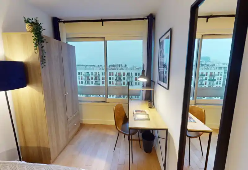 Bright, modern bedroom corner with a wooden wardrobe and a compact desk by a large window offering a city view. The image highlights usable workspace and warm wooden floors, ideal for listings focusing on comfort and natural light.