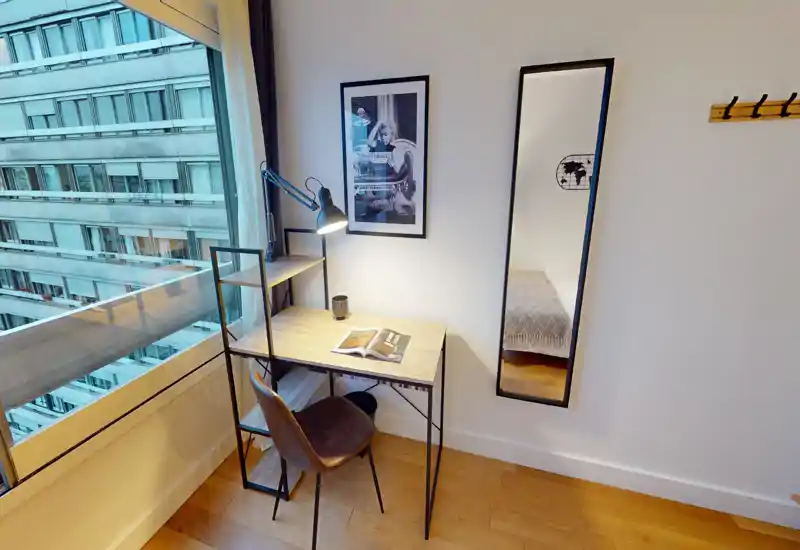 Compact bedroom corner with a small desk and chair beside a large window; a full-length mirror reflects the bed and adds depth. Clean modern styling with natural light and a reading lamp creates a bright, functional workspace inside the sleeping area.