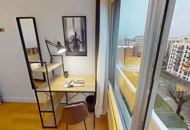 Coin chambre fonctionnel avec un bureau en bois compact et une lampe près d’une grande fenêtre offrant une vue sur la ville. Le lit est partiellement visible à gauche; un miroir et une affiche apportent du style.