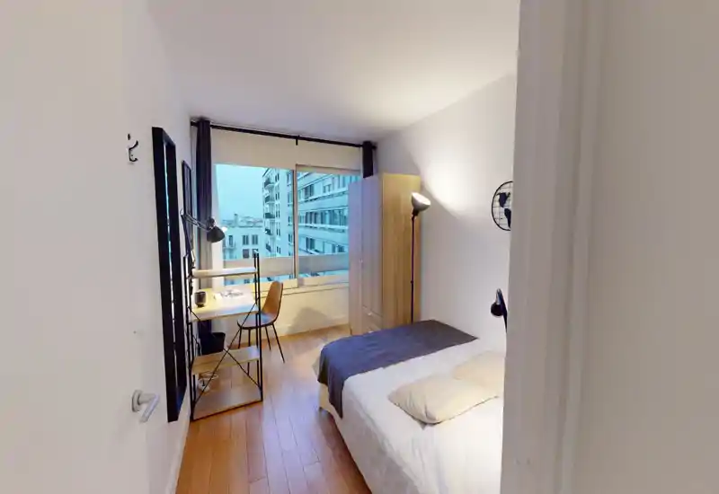 Chambre compacte et lumineuse avec un lit bien fait, parquet, armoire et un coin bureau près d'une grande fenêtre donnant sur la ville.