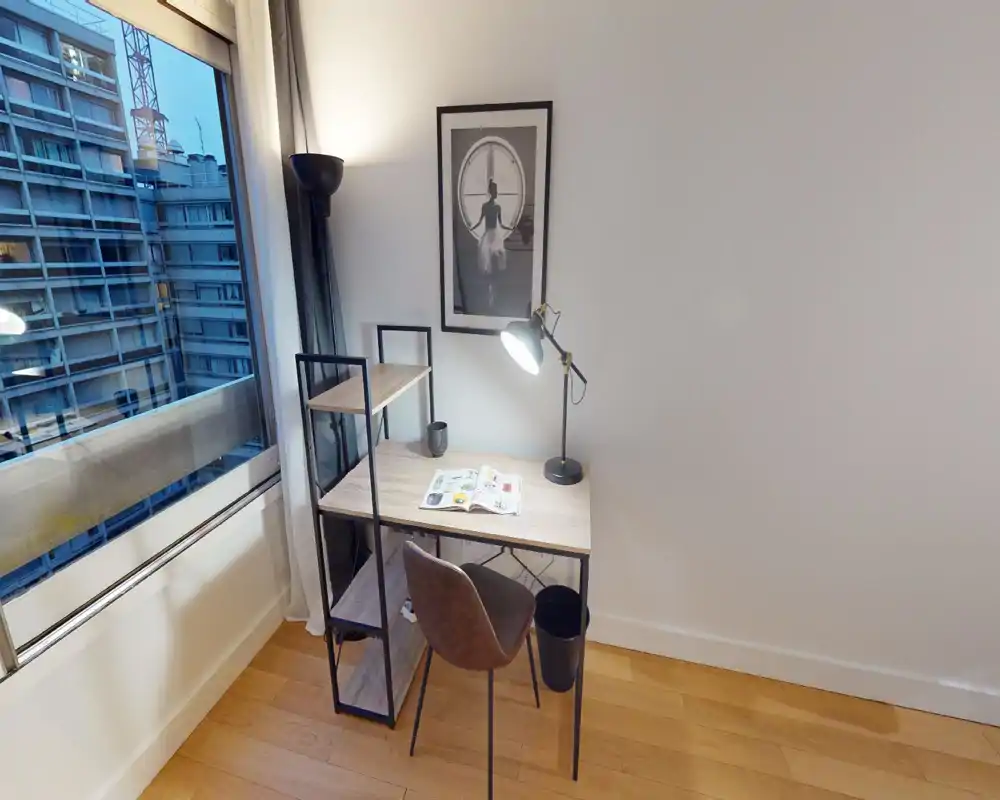 Angolo studio compatto nell'area living con scrivania in legno, lampada da lettura e finestra con vista sulla città — luminoso, ordinato e funzionale per il lavoro da remoto.