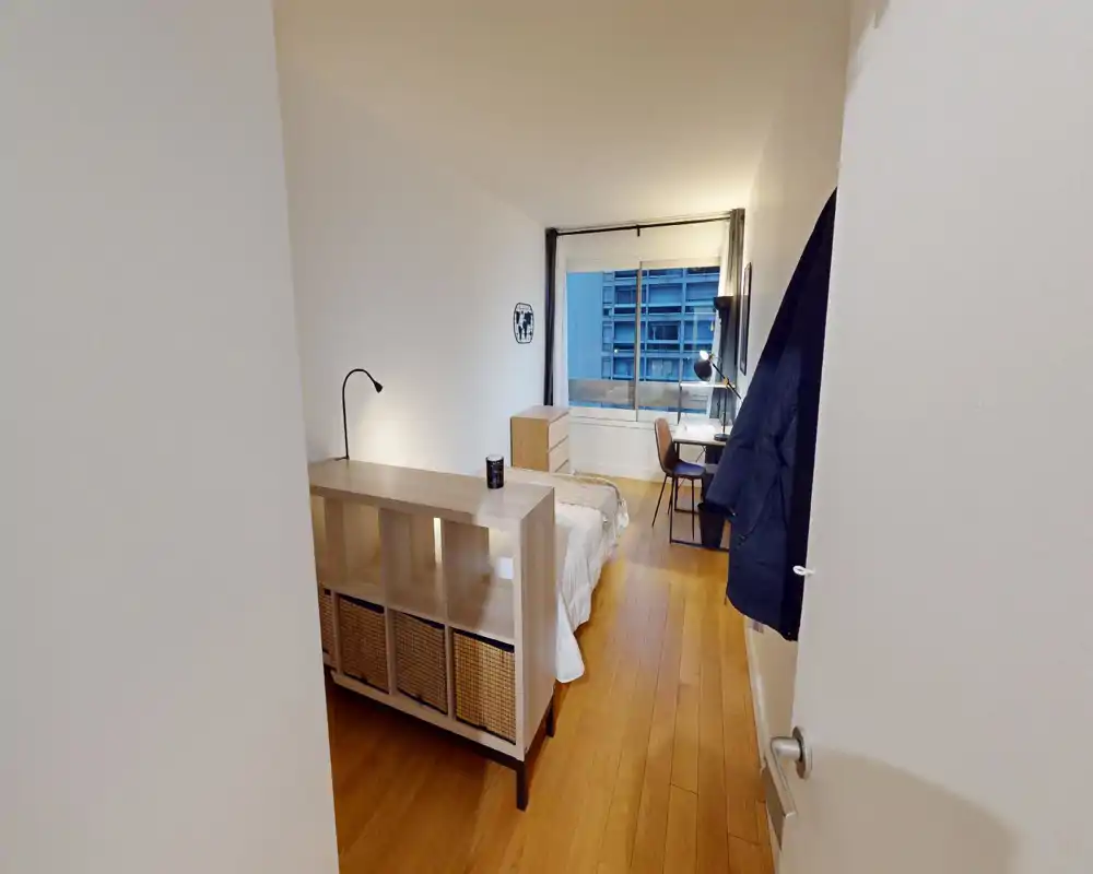 Camera da letto moderna e pulita con letto matrimoniale, scaffale in legno a piedi del letto, un piccolo comò e una scrivania vicino a una grande finestra con vista sulla città.