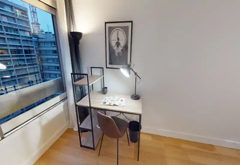 Angolo studio compatto nell'area living con scrivania in legno, lampada da lettura e finestra con vista sulla città — luminoso, ordinato e funzionale per il lavoro da remoto.