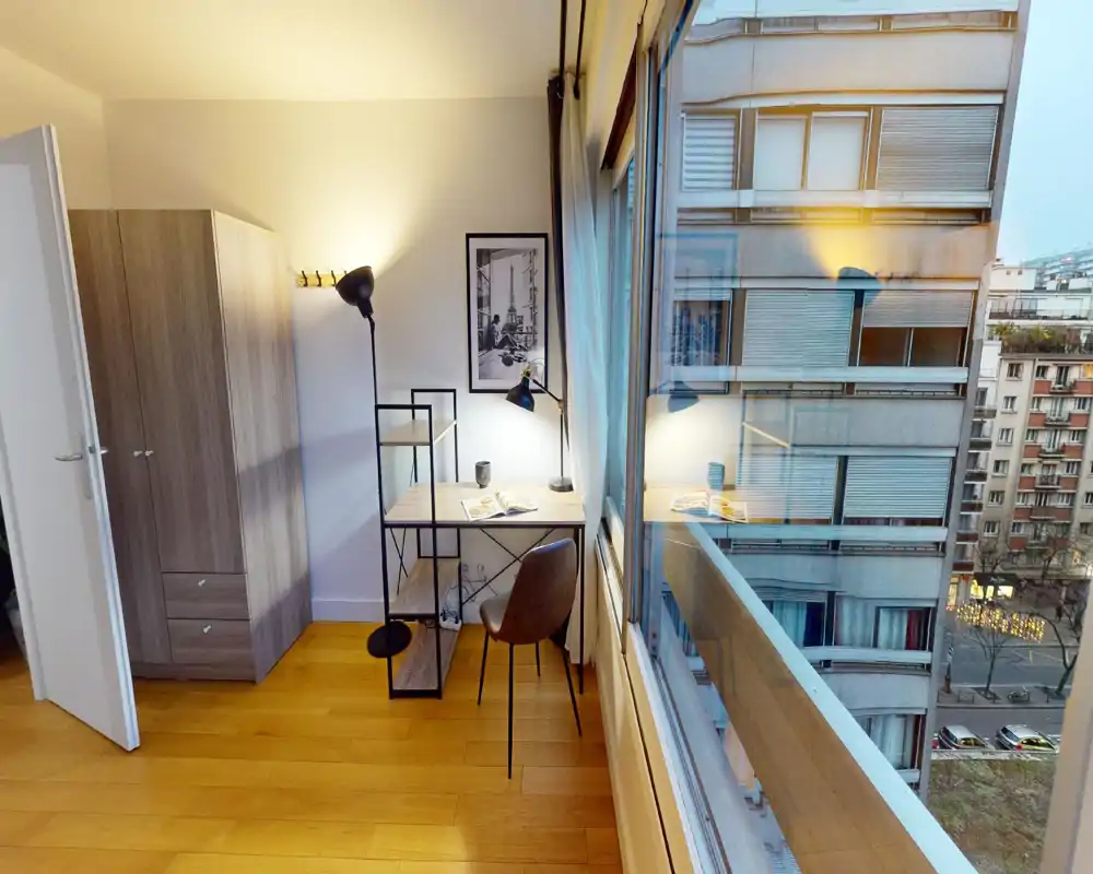 Coin de chambre lumineux et moderne avec bureau et armoire près d'une grande fenêtre donnant sur la rue. Sol en bois chaleureux, bureau avec lampe et chaise confortable.