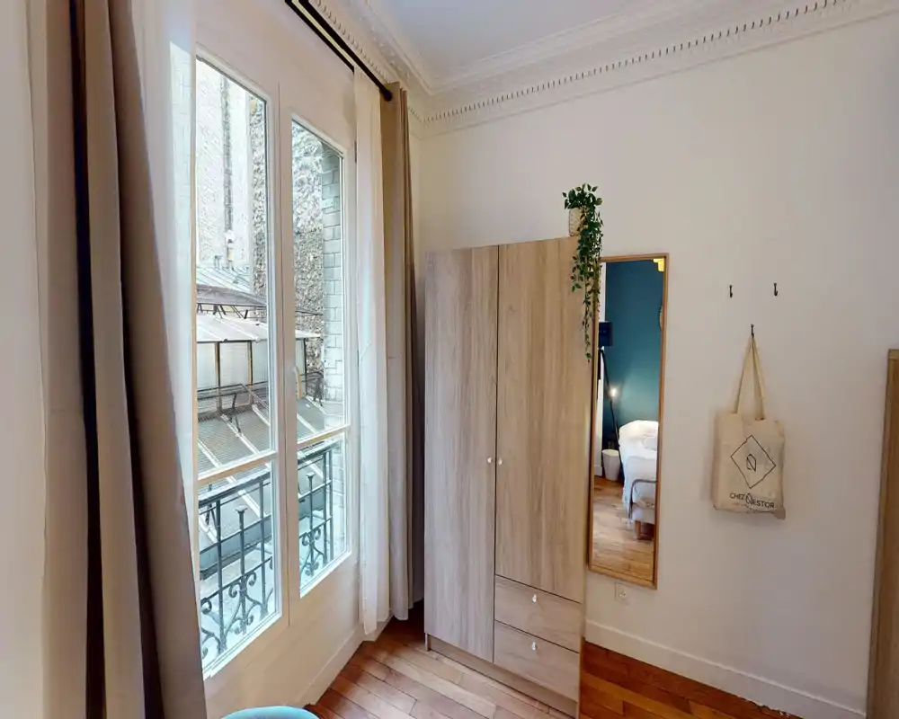Coin de chambre lumineux de style parisien avec une grande armoire en bois, un miroir pleine longueur et une grande fenêtre laissant entrer beaucoup de lumière naturelle.