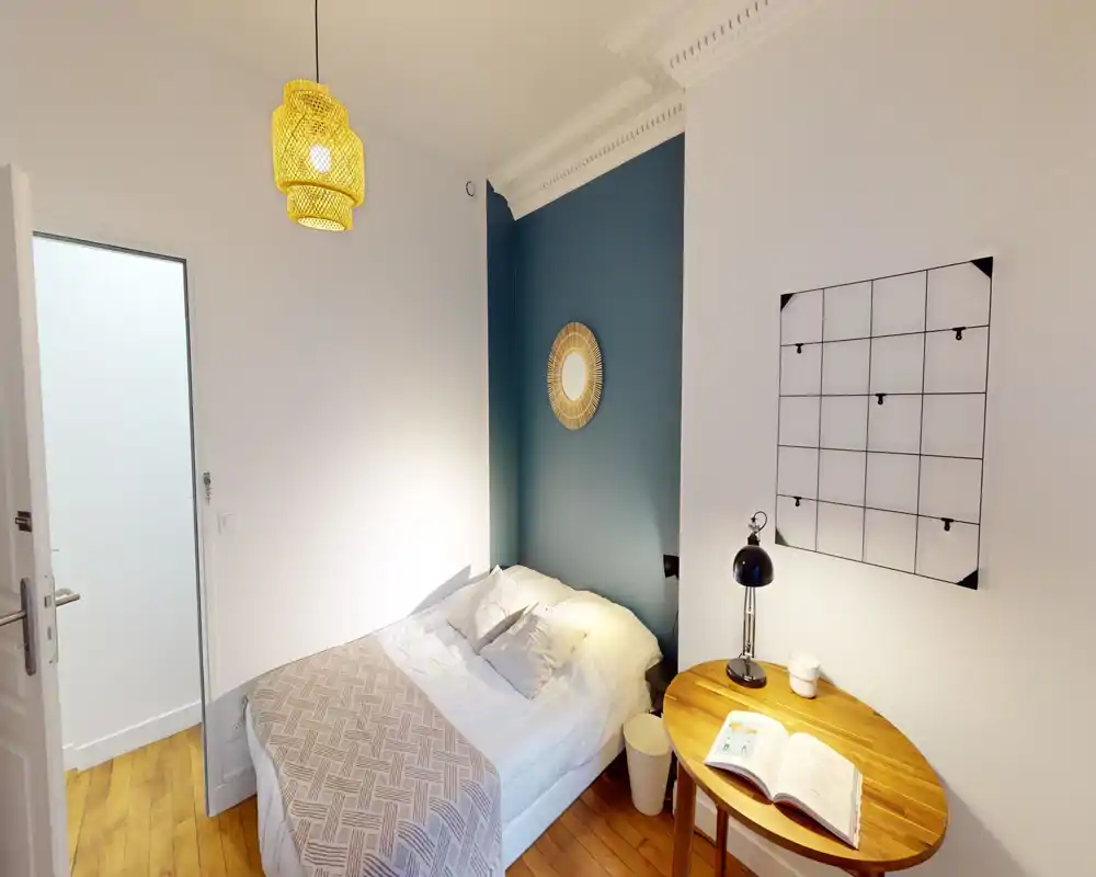 Chambre lumineuse et compacte avec un lit double contre un mur accentué bleu-vert, parquet chaud et petite table en bois avec lampe — accueillante et moderne.