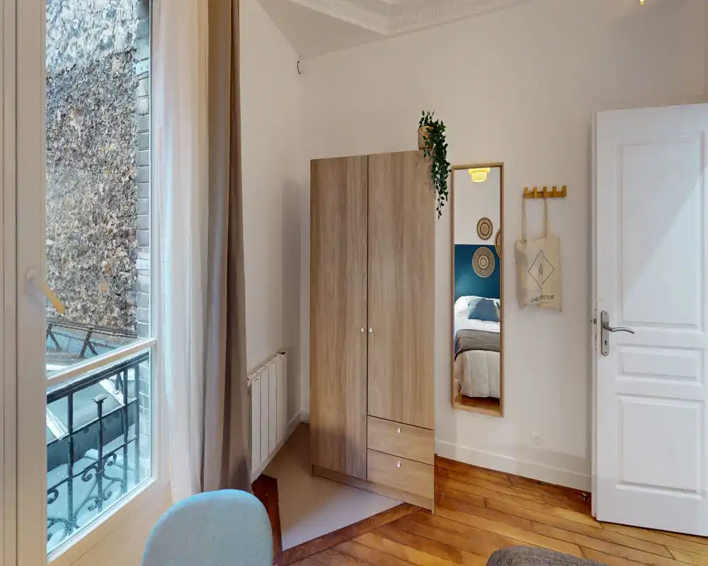 Coin de chambre lumineux avec une armoire en bois, un miroir en pied et un aperçu du lit reflété dans le miroir ; la lumière naturelle d’une grande fenêtre et le parquet chaleureux créent une ambiance accueillante.