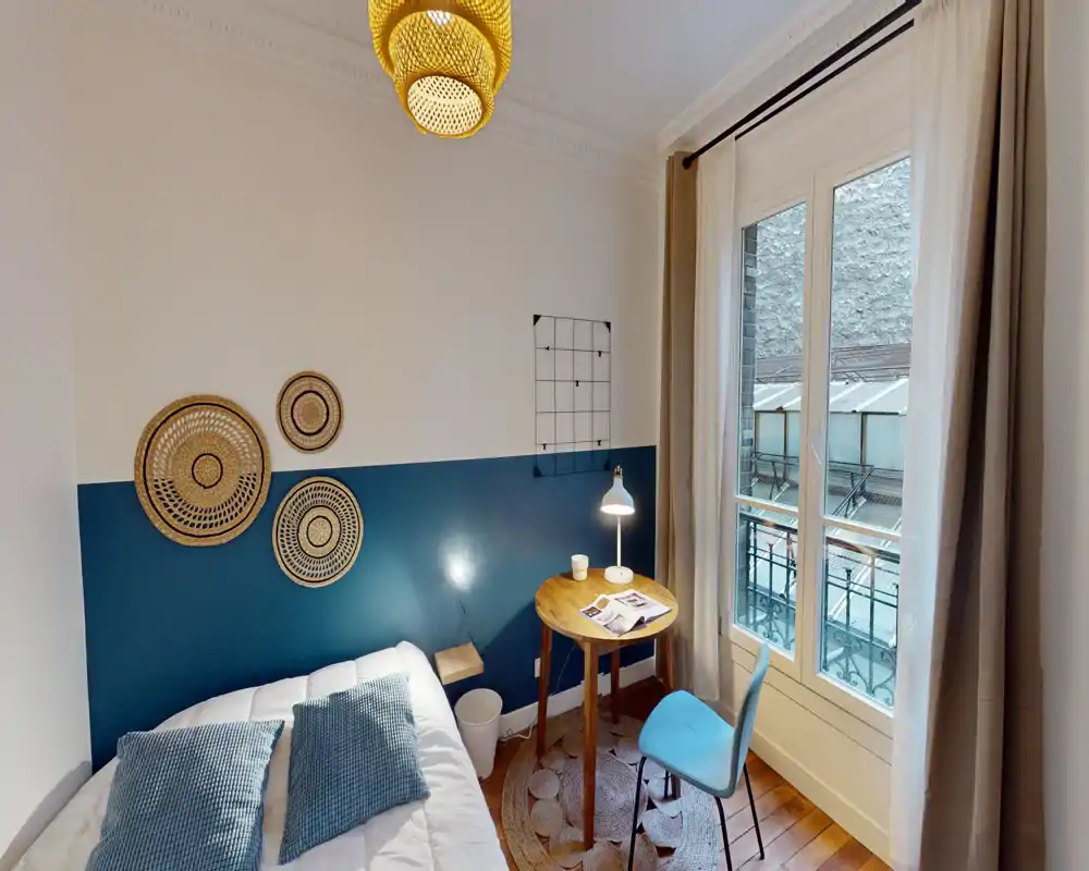 Chambre lumineuse et chaleureuse avec un lit double confortable, des décorations murales, une petite table ronde en bois avec lampe et une fenêtre laissant entrer la lumière naturelle — idéale pour un voyageur seul ou un couple.