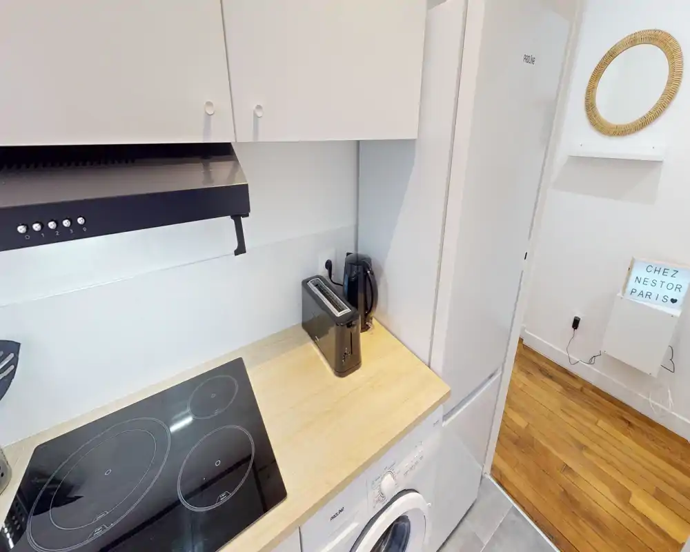Coin cuisine moderne et compact avec plaque à induction, plan de travail en bois, réfrigérateur et petits appareils (grille-pain, bouilloire), présenté dans un agencement lumineux et soigné.