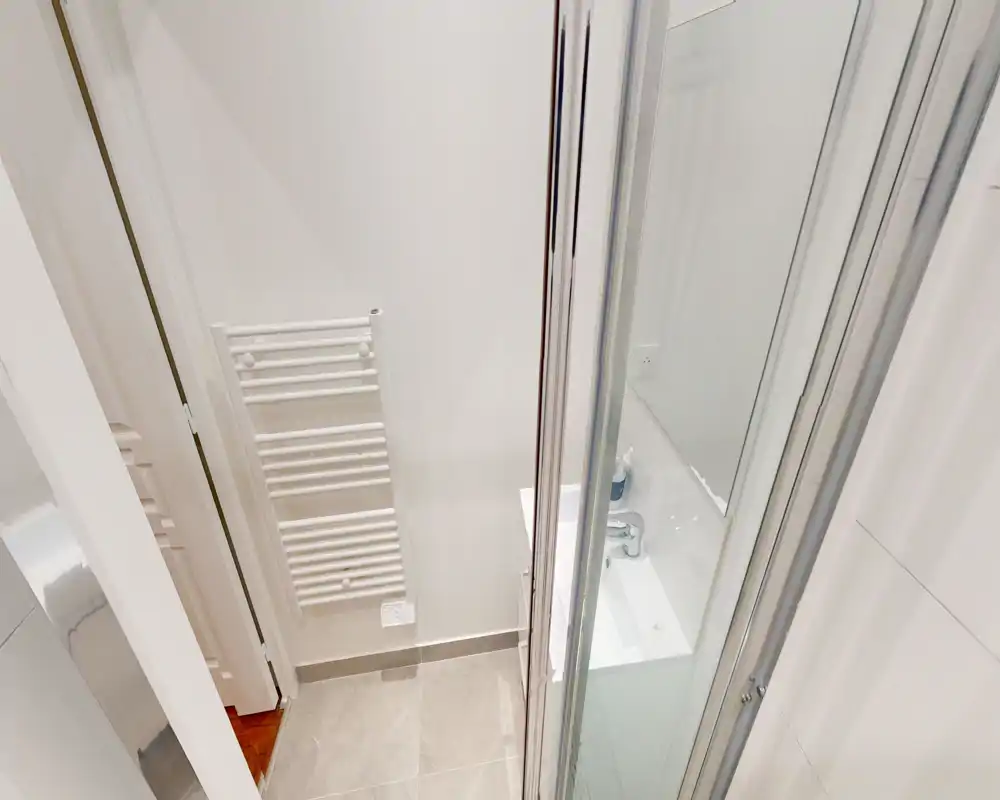 Salle de bain compacte et moderne avec sol carrelé, radiateur-serviettes mural, petit lavabo et porte de douche en verre visibles ; espace lumineux et propre adapté à une annonce.