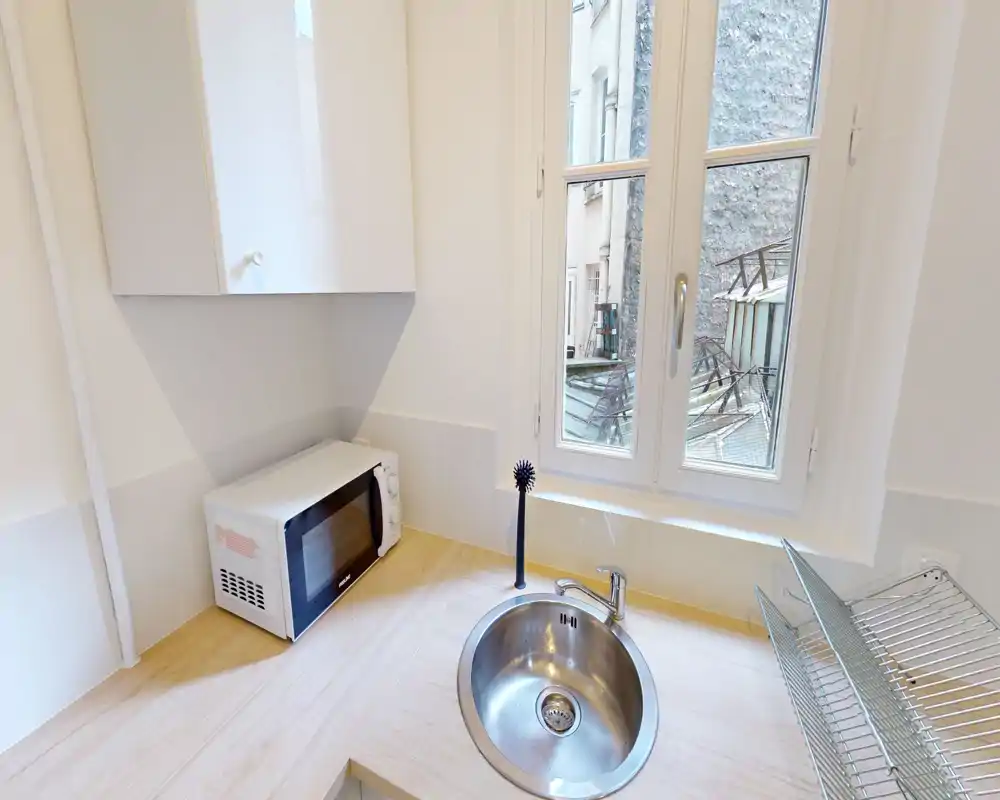 Kitchenette compacte avec un évier rond en acier inoxydable, un micro-ondes sur le plan de travail et une fenêtre apportant de la lumière naturelle ; agencement propre, minimal et fonctionnel, parfait pour des courts séjours.