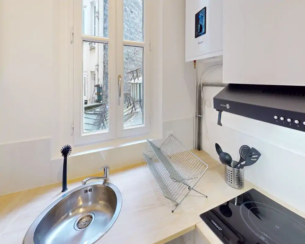 Kitchenette compacte et moderne avec évier en acier inoxydable, plaque à induction et égouttoir près d'une fenêtre lumineuse — propre et bien éclairée, parfaite pour les locations de courte durée.