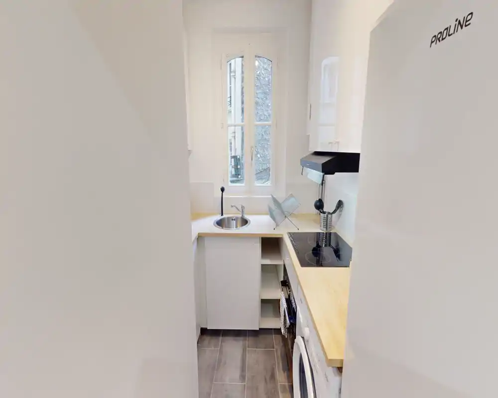 Kitchenette compacte et moderne avec plan de travail en bois clair, placards blancs et plaque induction à côté d'un évier rond sous une fenêtre haute apportant une bonne lumière naturelle.