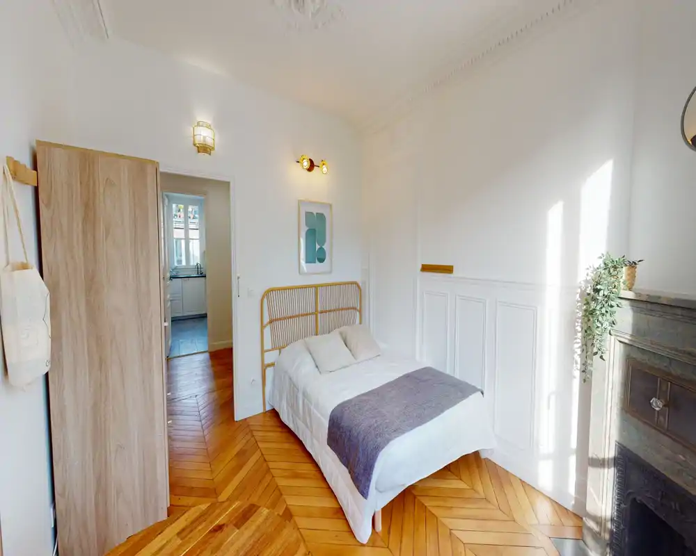 Chambre lumineuse et décorée avec un lit double, moulures élégantes et parquet en chevron chaleureux. La lumière naturelle inonde la pièce; une armoire et une cheminée apportent du charme.