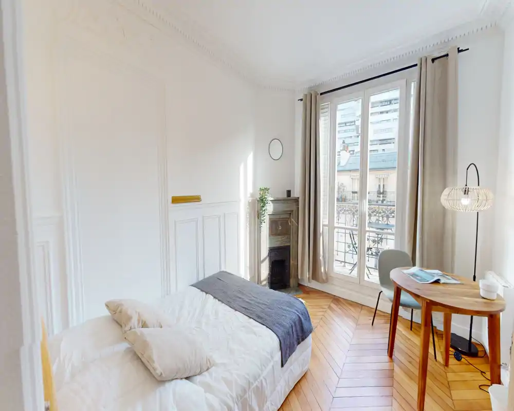 Chambre lumineuse et élégante avec un lit double confortable, hauts plafonds et grandes fenêtres à la française donnant sur un balcon. Parquet en chevron et textiles doux pour une atmosphère chaleureuse.