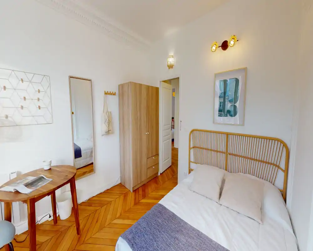 Chambre lumineuse et chaleureuse avec lit double à tête de lit en rotin, parquet chaleureux et mobilier en bois simple. Pièce bien éclairée aux murs neutres et décoration soignée, parfaite pour les locations courtes durées.