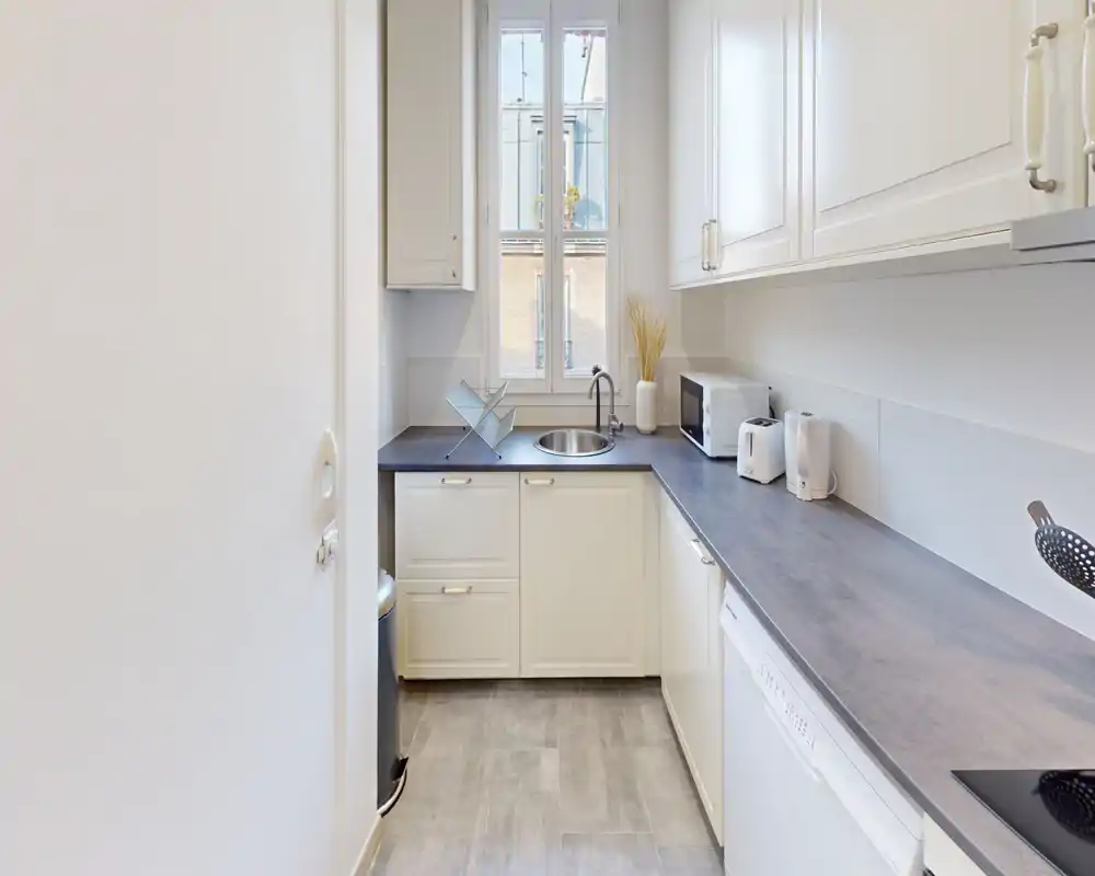 Cuisine en enfilade lumineuse avec placards blancs, plans de travail foncés et une fenêtre apportant de la lumière naturelle. Électroménagers intégrés et petits appareils sur le plan de travail, idéale pour un usage pratique en ville.