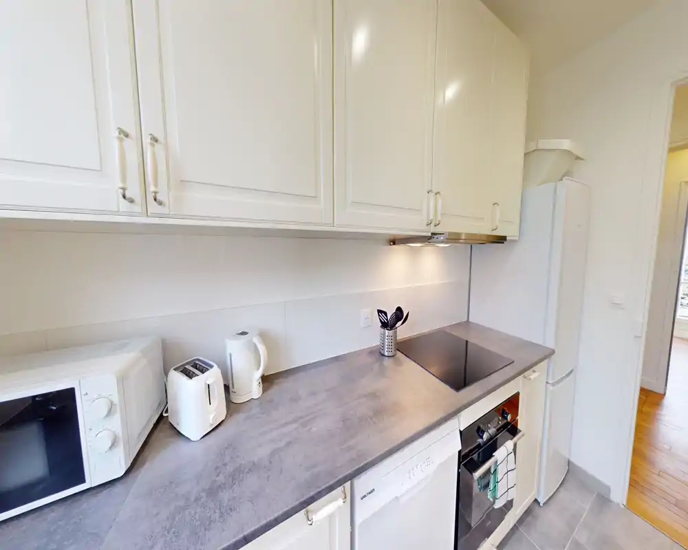 Cuisine moderne et lumineuse avec placards blancs, appareils intégrés, plaque à induction et micro-ondes/toaster sur le plan de travail — espace propre et fonctionnel pour cuisiner.