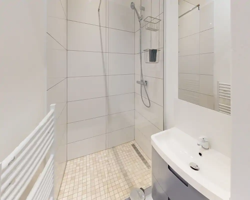 Salle de bain moderne et propre avec douche à l'italienne, sol carrelé et pommeau de douche mural. Petit lavabo et miroir visibles, finition blanche neutre.