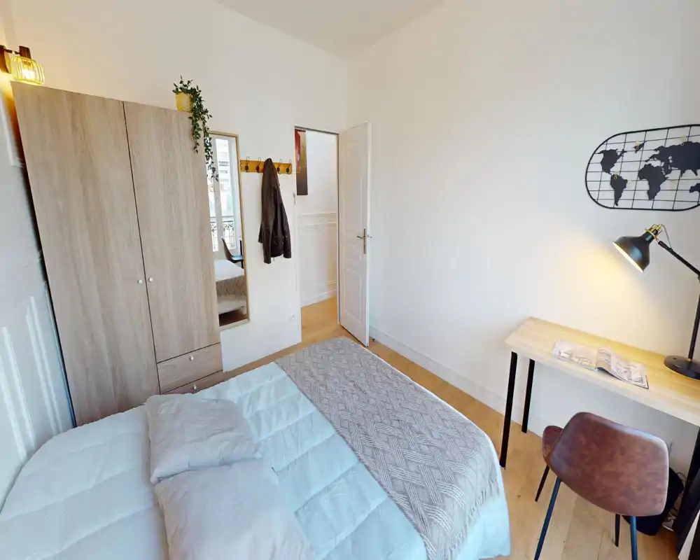Camera da letto luminosa e ordinata con un ampio letto in primo piano, un armadio e una scrivania con lampada — ideale per una persona o una coppia. Tessuti morbidi e una lampada calda creano un'atmosfera accogliente.