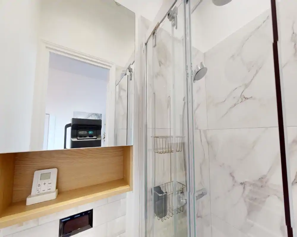 Bagno moderno con doccia in vetro, piastrelle effetto marmo e rubinetteria cromata; pensile specchiato e mensola in legno integrata per contenimento ed estetica.