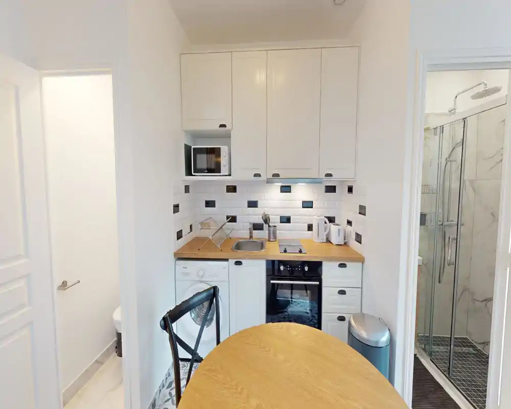 Cucina compatta e moderna con mobili bianchi, piano di lavoro in legno, paraschizzi piastrellato e elettrodomestici integrati tra cui forno, piano cottura, microonde e lavatrice. Luminosa e organizzata per un piccolo appartamento.