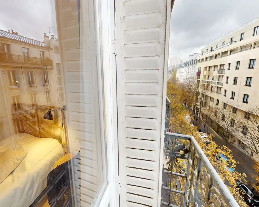 Camera da letto accogliente vista attraverso la finestra: letto con biancheria morbida, lampada da comodino e testata, con affaccio su una strada parigina dal balcone.
