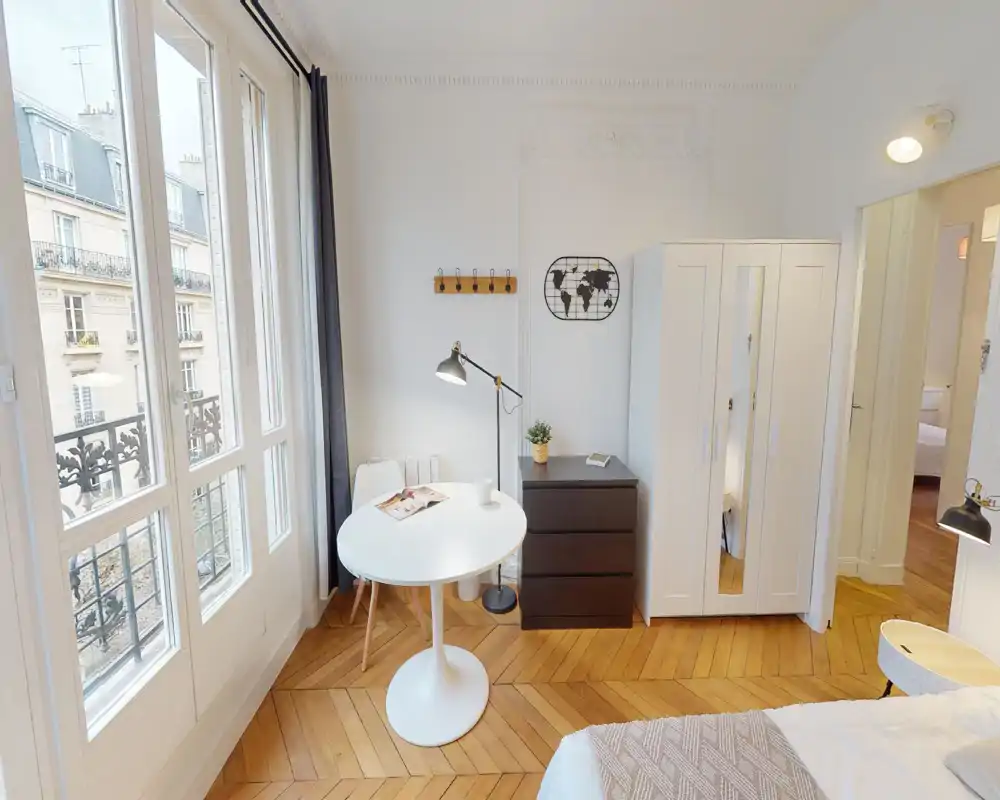 Camera luminosa e accogliente; camera da letto con ampie finestre, pavimento in parquet, armadio bianco e un piccolo tavolo per pranzo/lavoro accanto al letto.