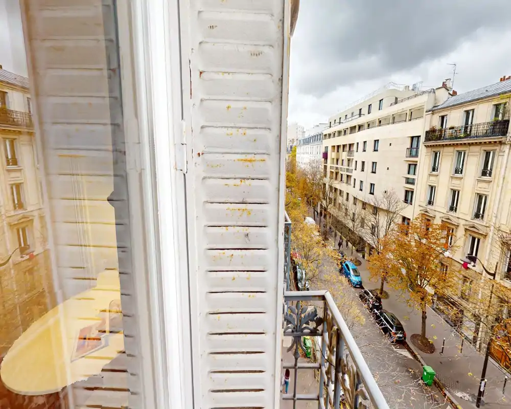 Pequeño balcón con vistas a una calle arbolada de estilo parisino con fachadas clásicas y coches aparcados; barandilla de hierro forjado decorativa y contraventana enmarcan la escena.