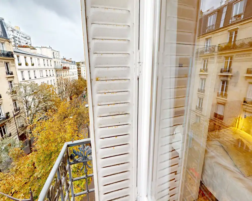 Vista desde una ventana de estilo parisino con un pequeño balcón y árboles otoñales, con una parte de un dormitorio acogedor reflejada en el vidrio.