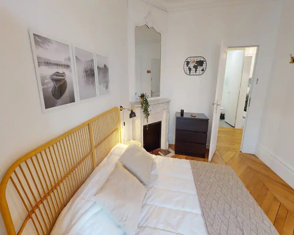 Dormitorio luminoso y con estilo, con cabecero de ratán, ropa de cama blanca y una chimenea de mármol decorativa; suelos de madera cálida y cuadros en la pared añaden confort.