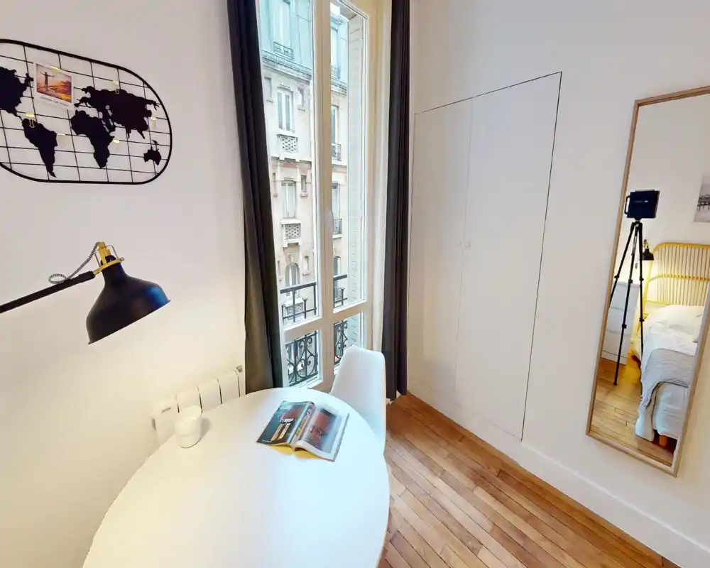 Rincón de dormitorio luminoso y de estilo sencillo con ventana y armario. Una mesa redonda y una silla crean un pequeño espacio de trabajo; la cama y la lámpara de noche se ven parcialmente en el espejo, indicando un espacio para dormir compacto y acogedor.