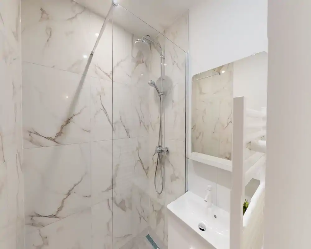 Bagno moderno e compatto con doccia walk-in e soffione a pioggia, piastrelle effetto marmo e piccolo lavabo con specchio — luminoso e pulito.