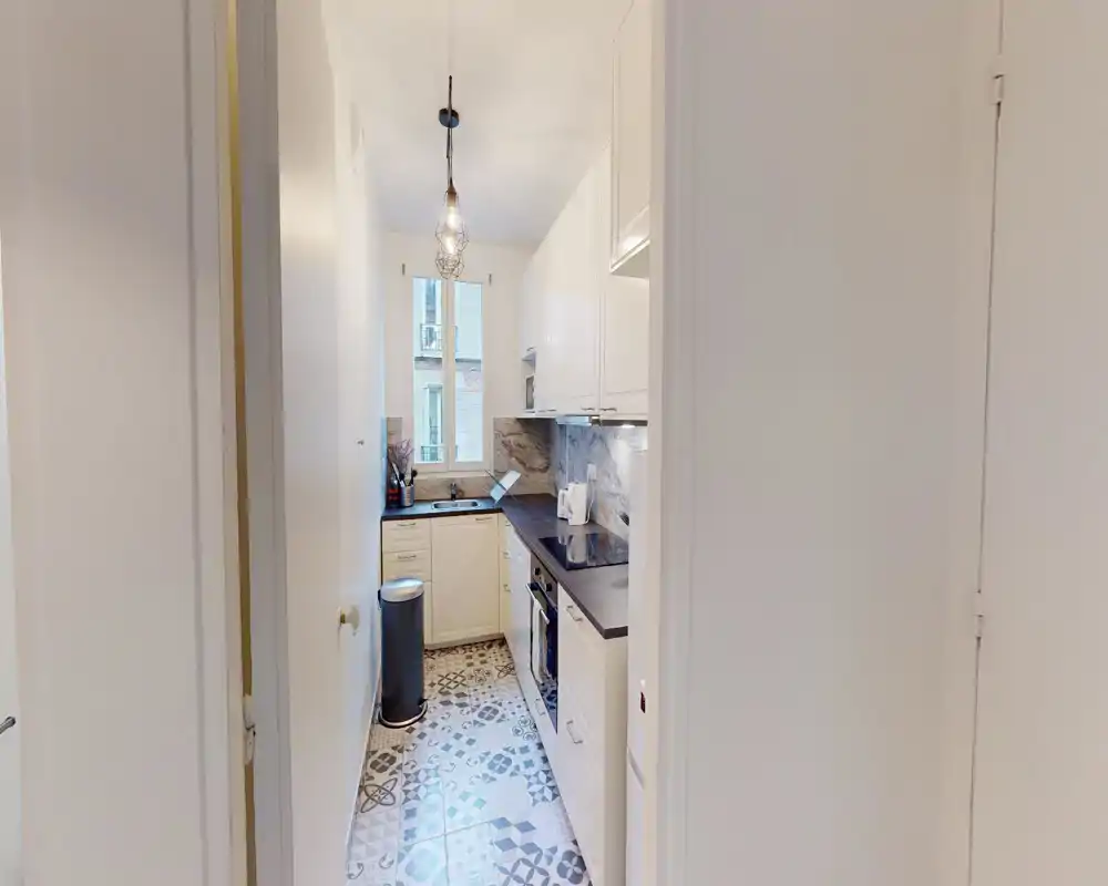 Cucina stretta e luminosa con pensili bianchi, piano scuro e piastrelle a motivi decorativi; lampada a sospensione e finestra rendono l’ambiente accogliente e ben illuminato.