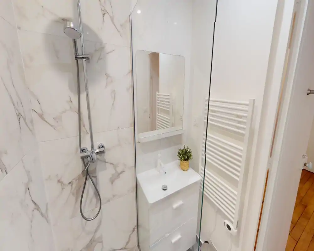 Bagno moderno e luminoso con doccia a filo, piastrelle effetto marmo bianche, lavabo compatto con specchio e termoarredo.