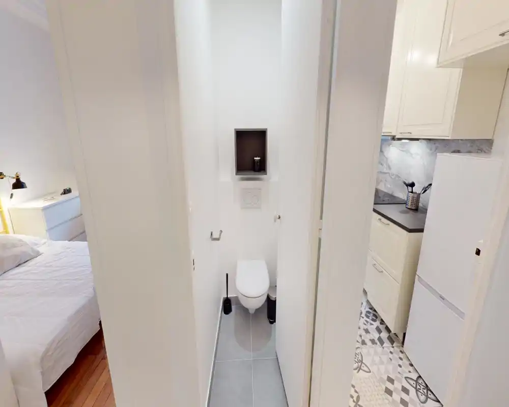 Piccolo bagno con WC sospeso, piastrelle grigio chiaro e pareti bianche; camera da letto e cucina adiacenti sono visibili attraverso le porte.