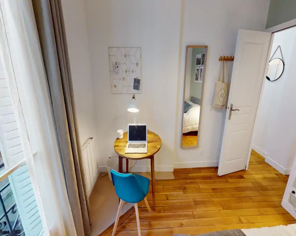 Coin de chambre compact avec un petit bureau en bois près d'une grande fenêtre, une chaise bleue et un aperçu du lit visible au bord de l'image et dans le miroir mural. Sol en bois chaleureux, lumière naturelle douce et touches décoratives simples pour une atmosphère cosy et lumineuse.