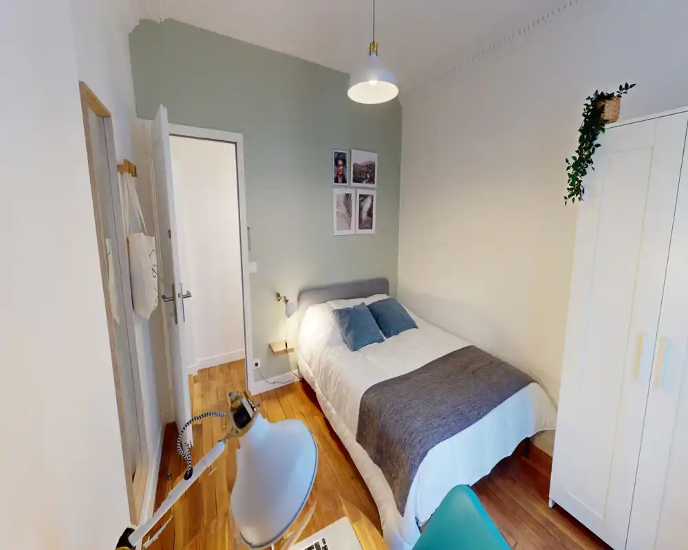 Chambre soignée avec lit confortable, coussins moelleux et plaid gris. Sol en bois clair et mur d'accent vert pâle apportent une atmosphère calme et bien mise en scène, parfaite pour une annonce locative.