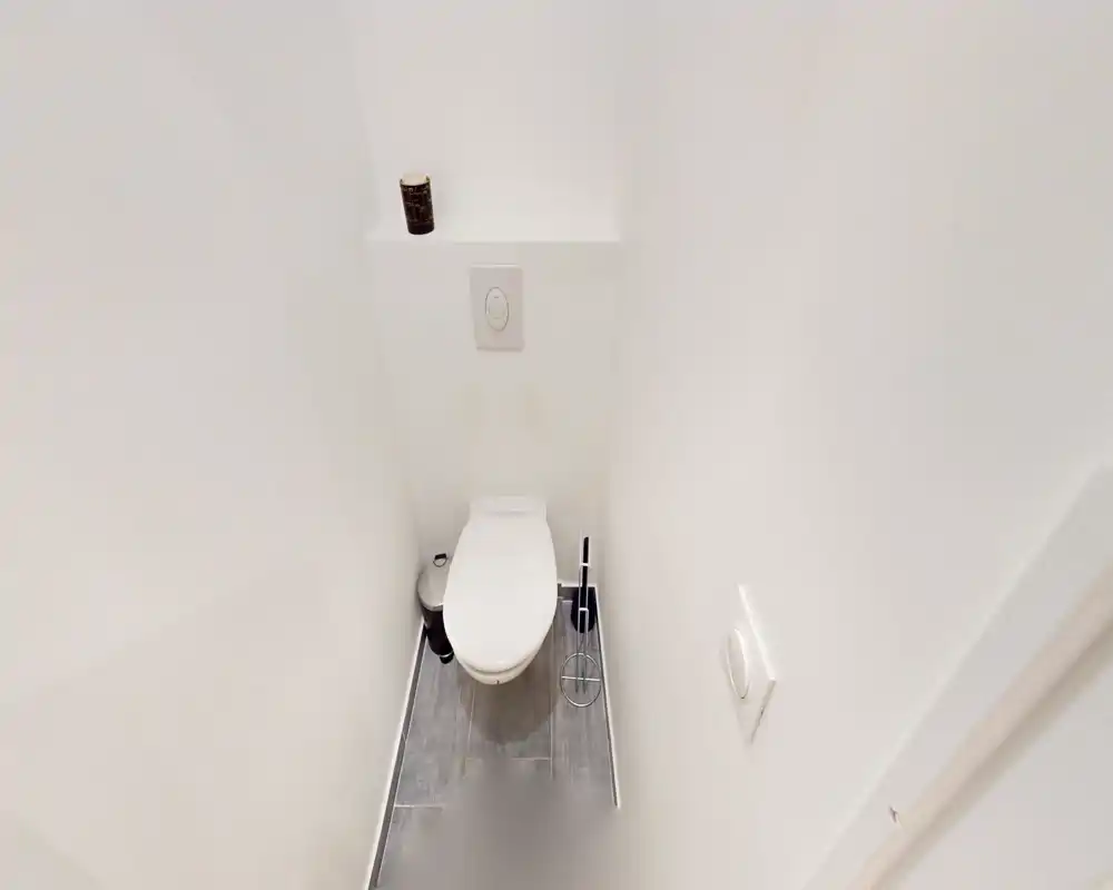 WC compact et moderne avec plaque de chasse d'eau murale et sol carrelé. Murs blancs et accessoires minimalistes pour des toilettes d'appoint propres et faciles à entretenir.