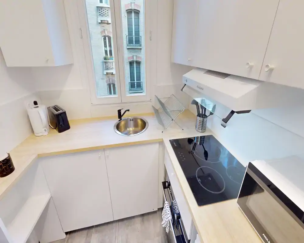 Cuisine moderne et compacte avec plan de travail en bois clair, plaque à induction, four et micro-ondes encastrés. Pièce lumineuse et bien rangée avec évier sous la fenêtre et petits appareils sur le plan de travail.