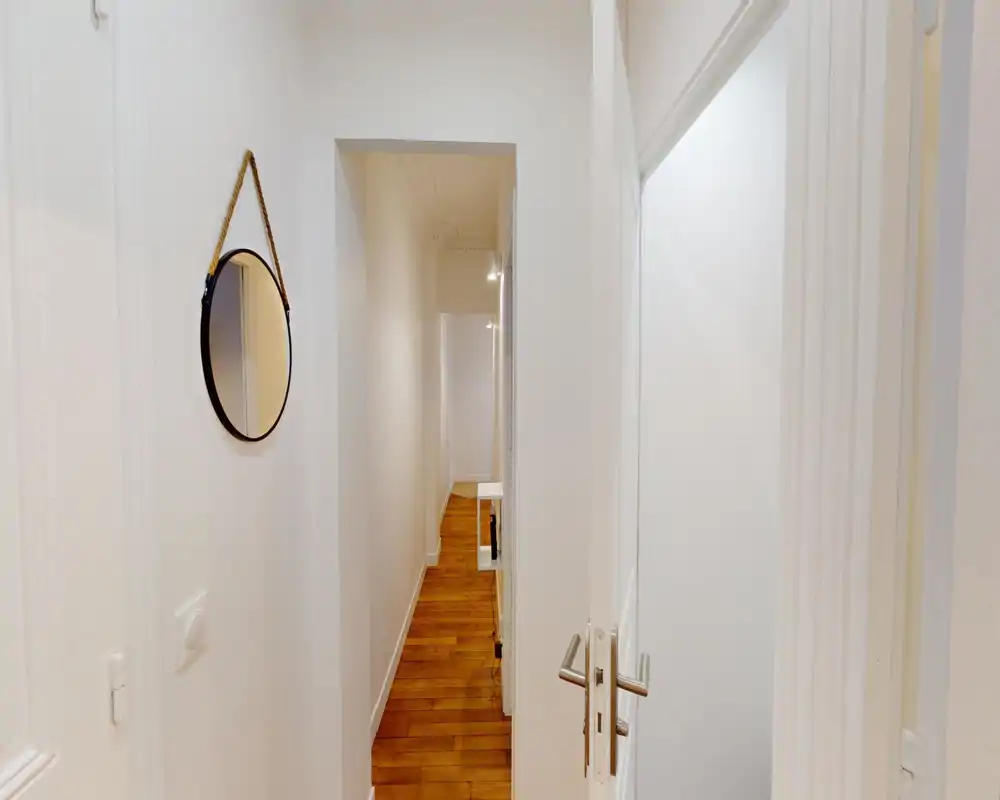 Couloir d'entrée étroit aux murs blancs, parquet en bois et miroir rond accroché au mur ; lumineux, minimaliste et bien entretenu.
