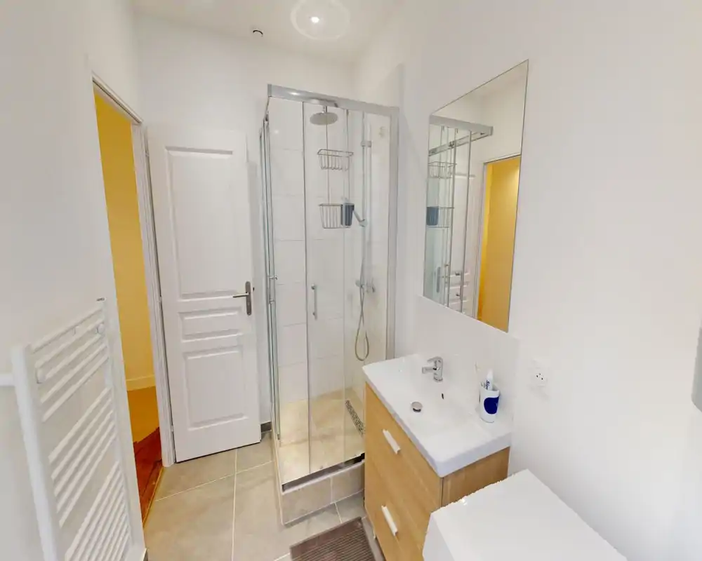 Salle de bains moderne et compacte avec douche vitrée, meuble lavabo en bois, grand miroir et sèche-serviettes chauffant. Murs blancs lumineux et éléments contemporains pour une impression nette et fraîche.