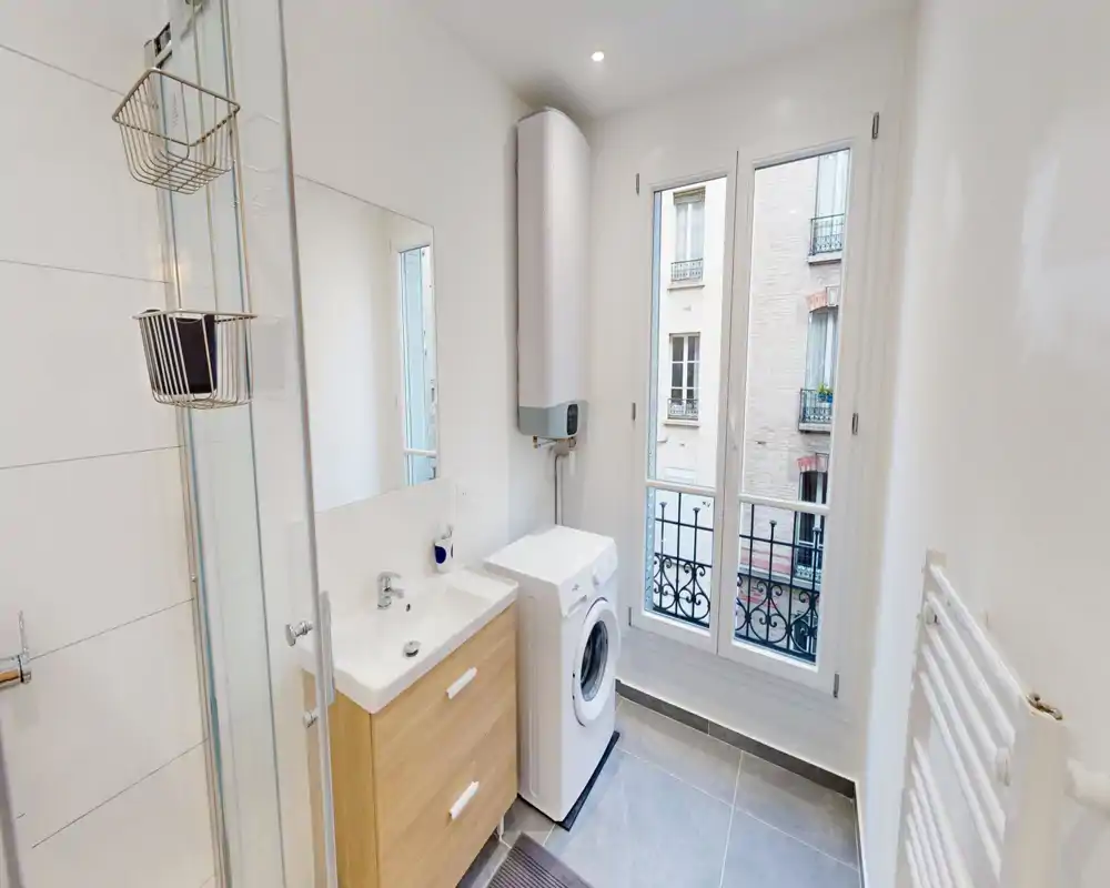 Salle de bains moderne et lumineuse avec douche, meuble vasque et machine à laver près d'une grande fenêtre apportant une belle lumière naturelle.
