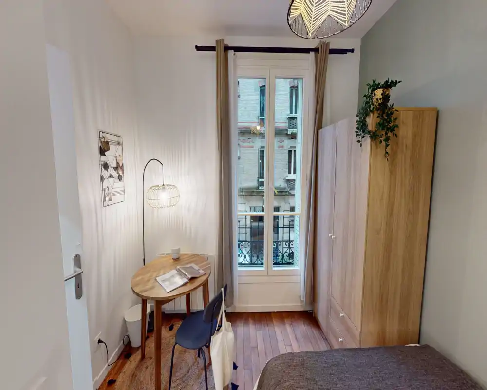 Camera da letto accogliente e ben illuminata con pavimenti in legno, letto compatto, armadio e un piccolo tavolo rotondo accanto a una finestra alta che porta luce naturale.