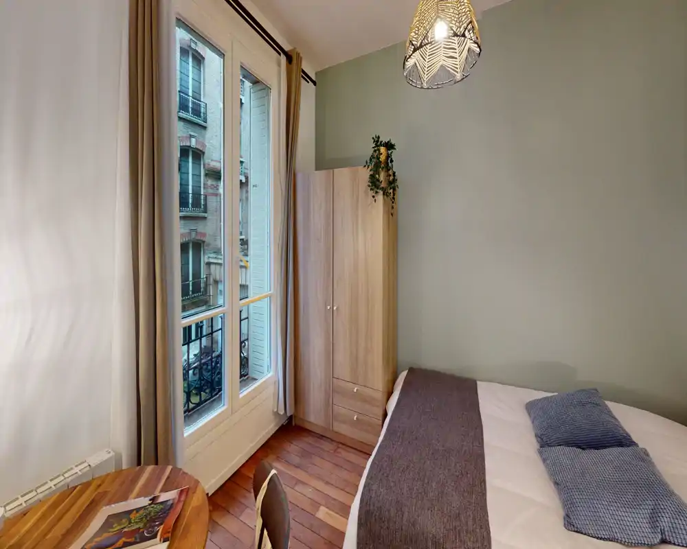 Camera da letto compatta e luminosa con letto matrimoniale, tessili accoglienti e un armadio in legno. La grande finestra illumina la stanza e il pavimento in legno caldo con il lampadario decorativo dona carattere.
